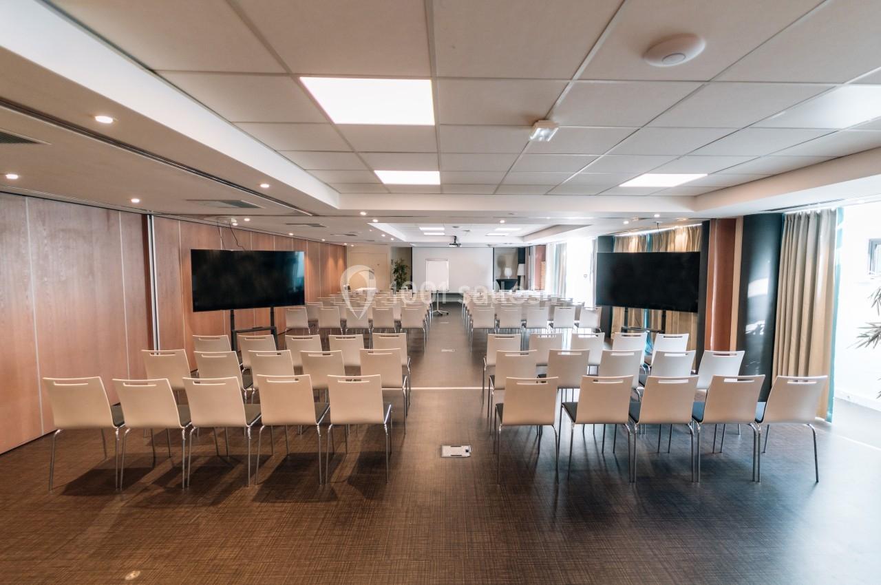 Salle de conférence lumineuse avec des rangées de chaises blanches, deux écrans et un projecteur au fond.