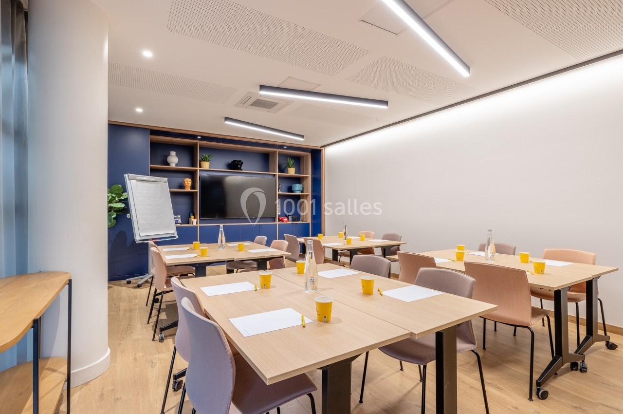 Salle de réunion lumineuse avec tables, chaises, tableau de conférence, écran et fournitures disposées pour un atelier.