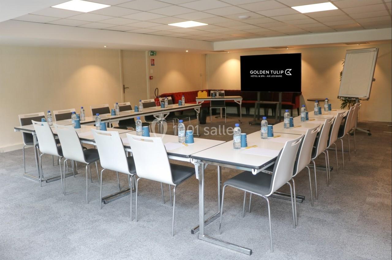Salle de réunion lumineuse avec tables disposées en U, chaises blanches, écran de présentation et tableau à feuilles.