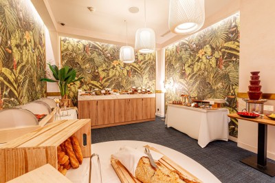 Buffet de petit-déjeuner avec viennoiseries, pain, fruits et fontaine de chocolat dans une salle décorée de motifs tropicaux.