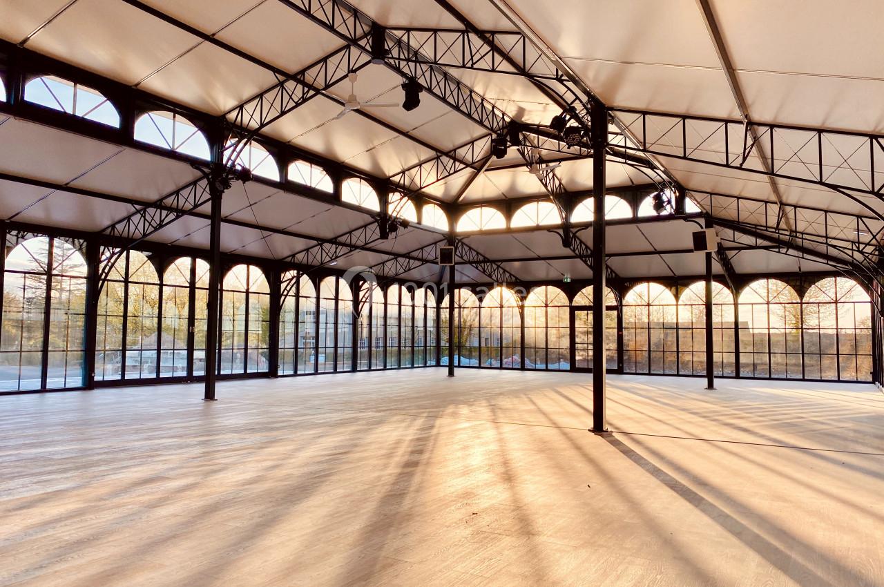 Grande salle lumineuse avec structure métallique, toit en toile blanche et larges baies vitrées laissant entrer la lumière…
