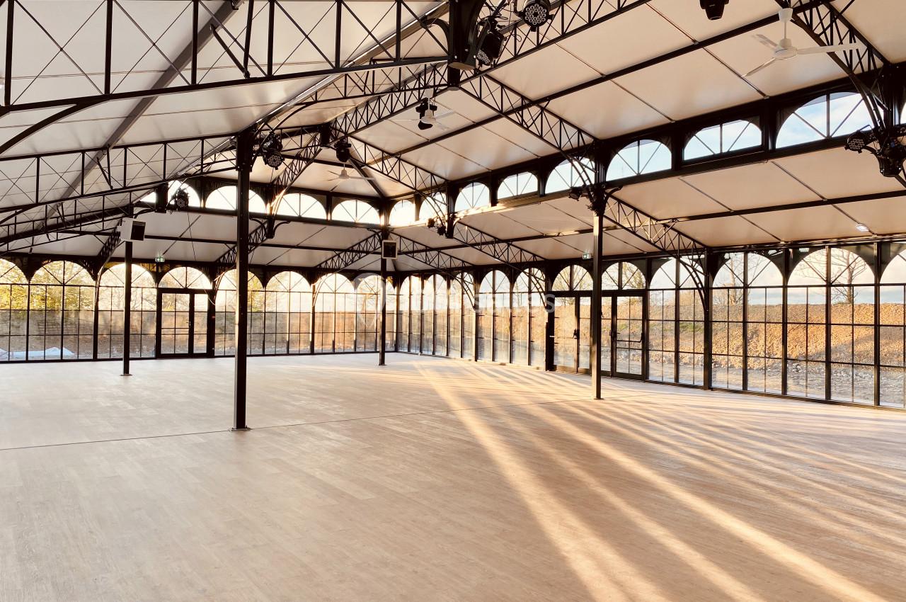 Intérieur d'une grande salle lumineuse avec structure métallique, toit en verre et sol en bois clair.