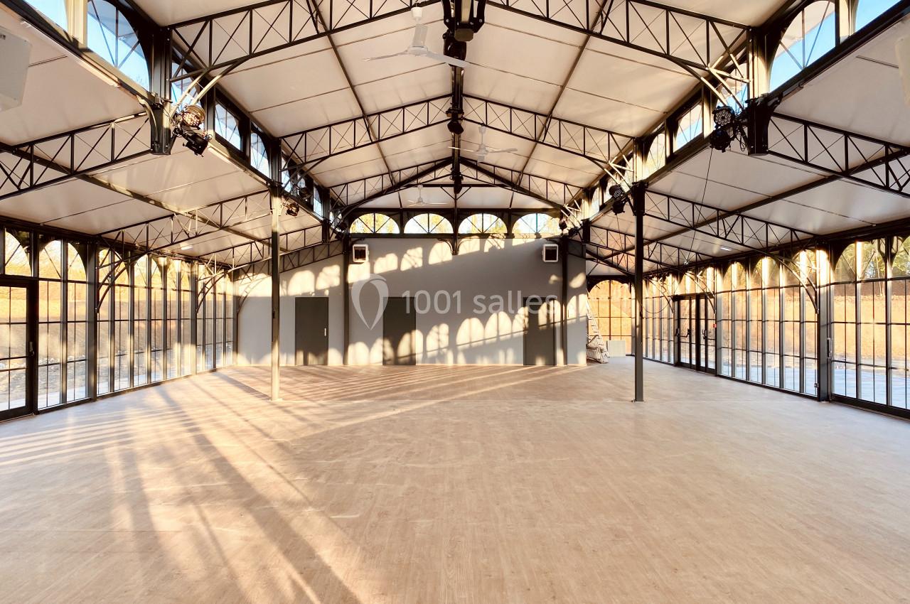Grande salle lumineuse avec structure métallique, murs vitrés et sol en bois clair, baignée par la lumière du soleil.