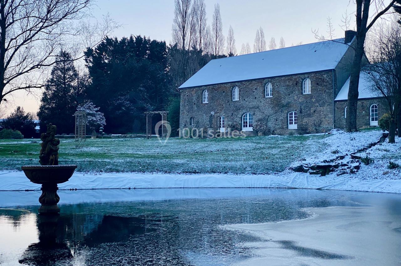 Manoir en pierre entouré d'un jardin enneigé avec un bassin gelé et une fontaine au premier plan.