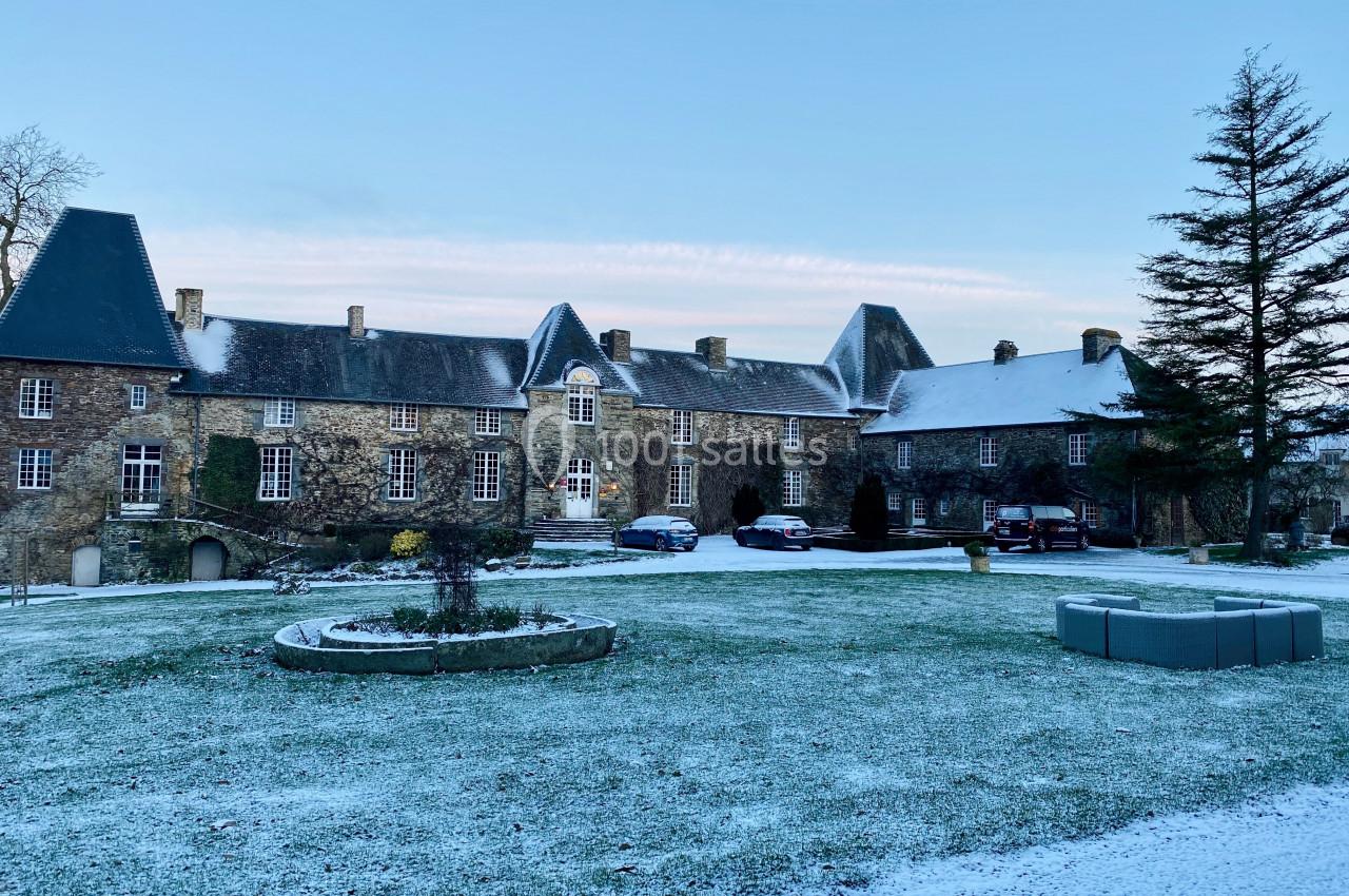 Façade d'un bâtiment en pierre avec toits en ardoise, entouré d'une pelouse légèrement enneigée et d'arbres.