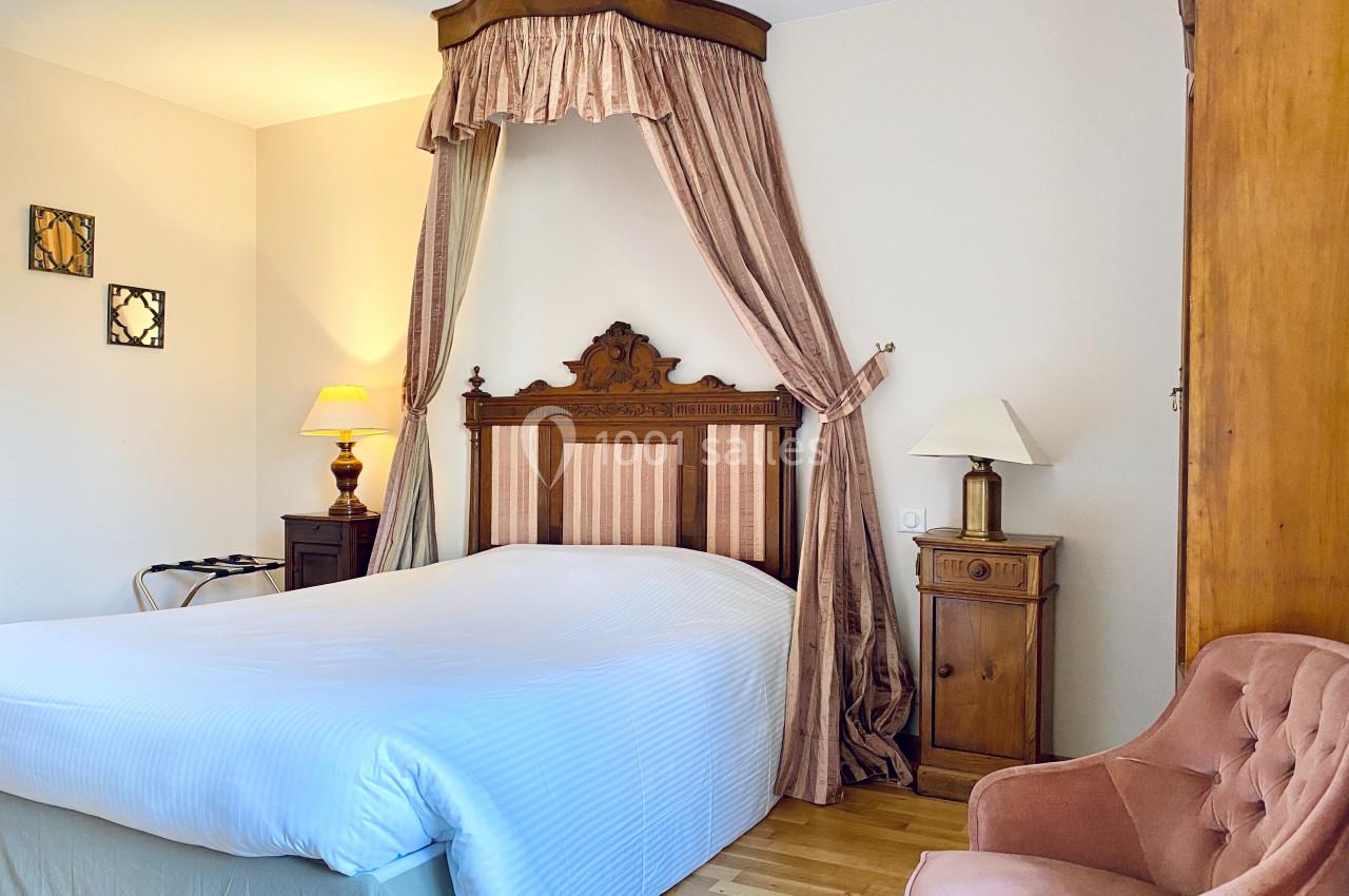 Lit double avec baldaquin en bois sculpté, draps blancs, fauteuil rose et lampes de chevet dans une chambre lumineuse.