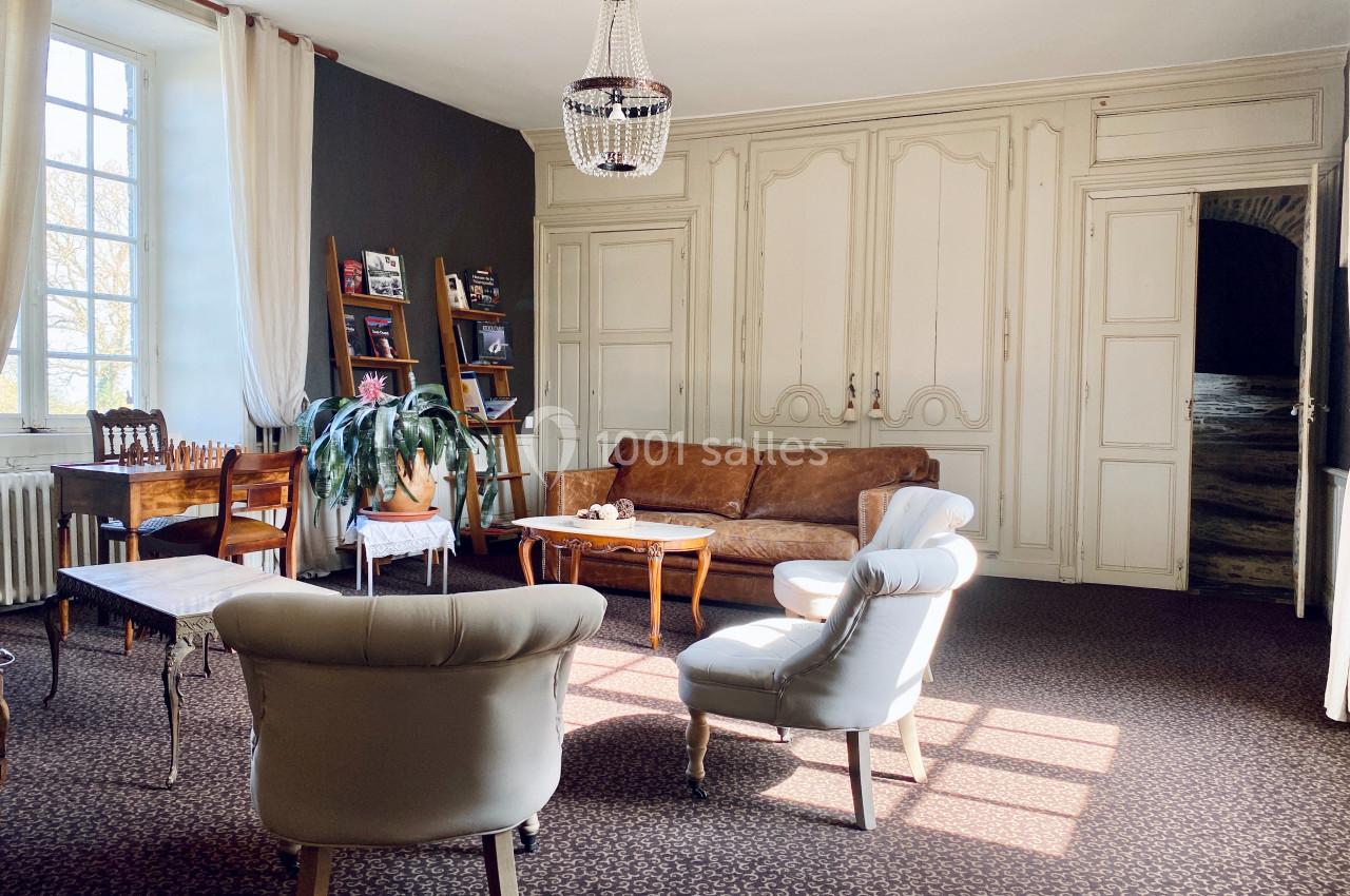 Salon lumineux avec canapé en cuir, fauteuils blancs, table basse en bois, étagères et grande fenêtre.