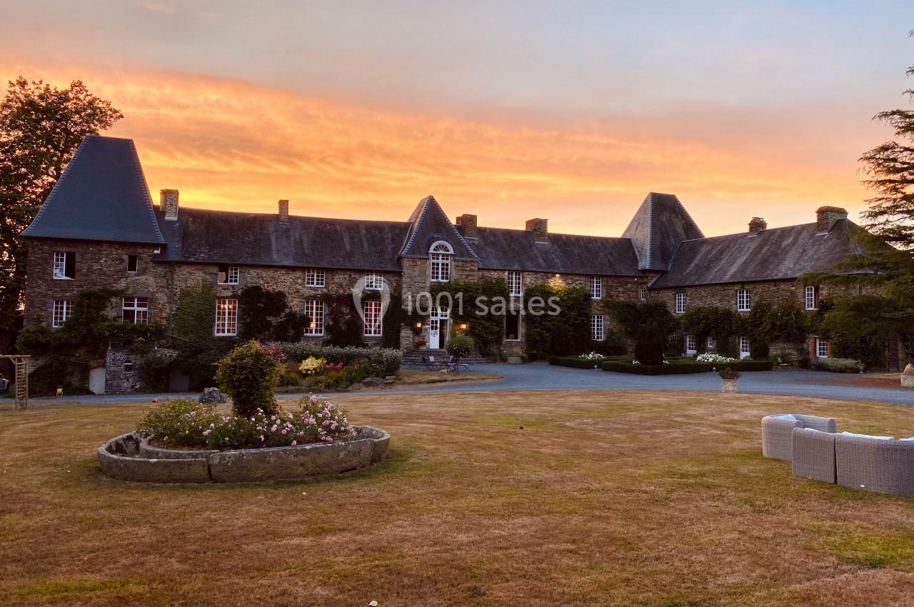 Manoir en pierre avec toit en ardoise, entouré de pelouse et de fleurs, sous un ciel de coucher de soleil.