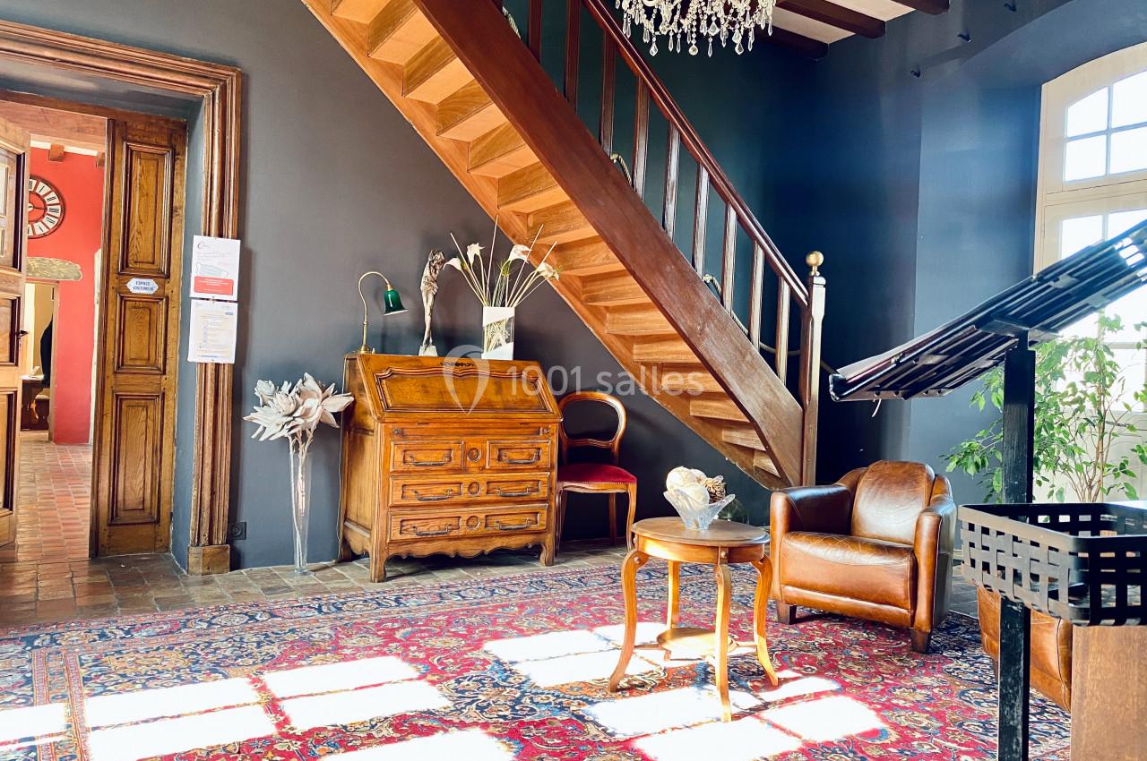 Hall lumineux avec escalier en bois, tapis coloré, mobilier ancien, fauteuil en cuir et lustre suspendu.