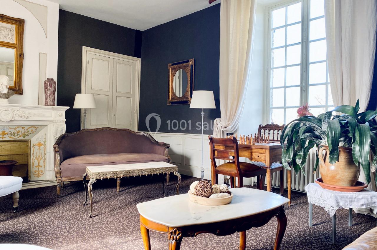 Salon lumineux avec canapé, table basse, cheminée décorative, plantes et mobilier ancien près d'une grande fenêtre.