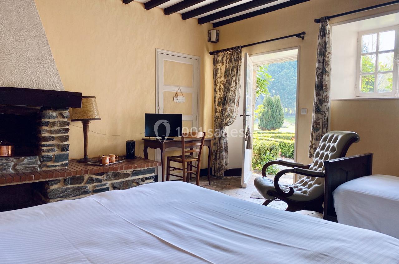 Chambre lumineuse avec lit, cheminée en pierre, bureau, fauteuil et vue sur un jardin à travers une porte ouverte.