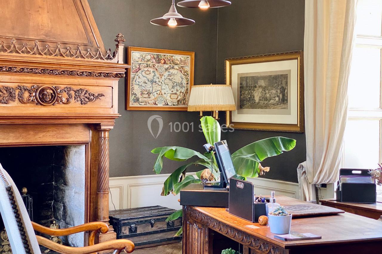Bureau élégant avec cheminée en bois sculpté, plantes, cartes encadrées et lumière naturelle entrant par une fenêtre.