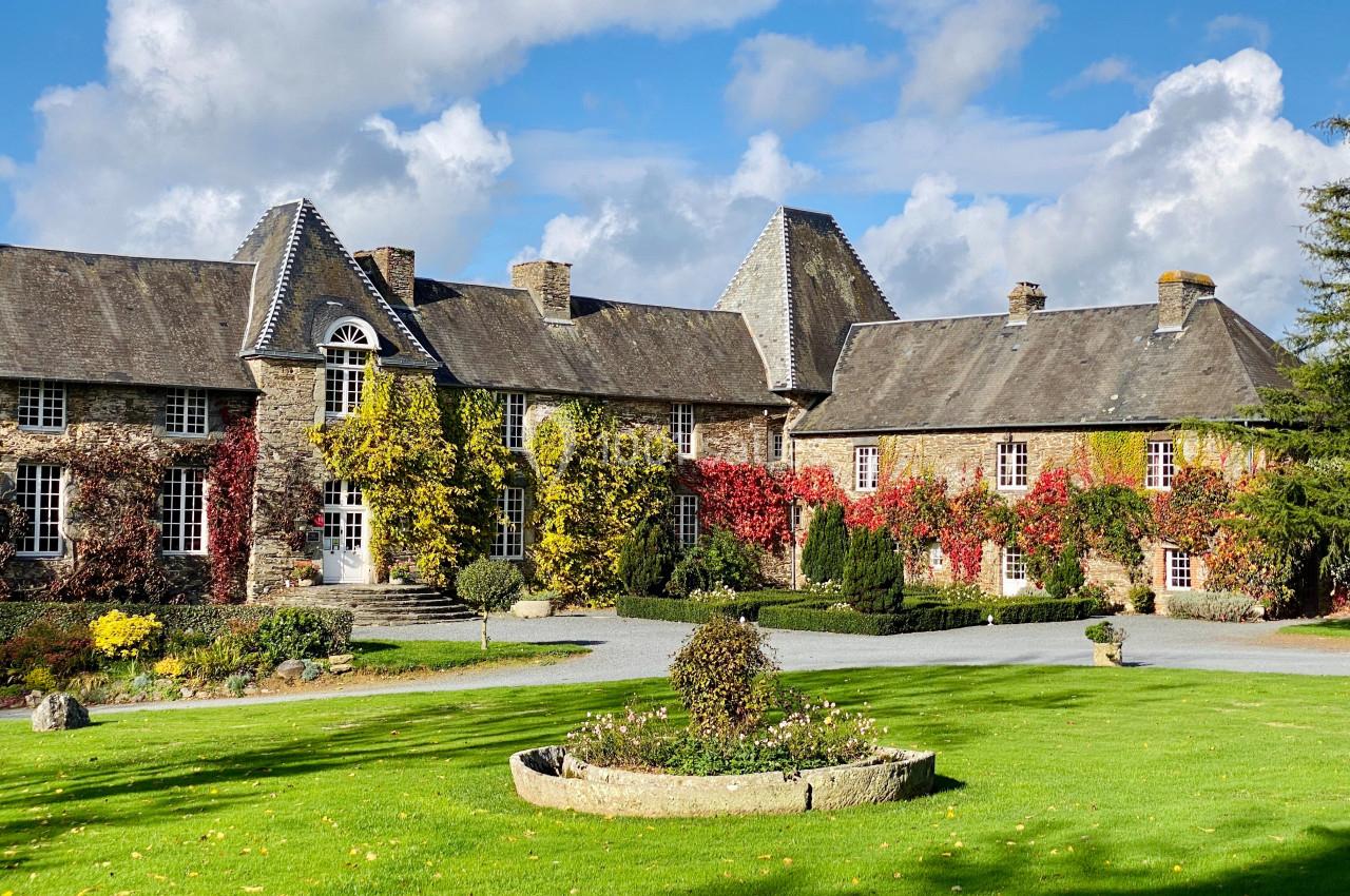 Manoir en pierre entouré de vignes rouges et jaunes, avec pelouse et parterre fleuri au premier plan.