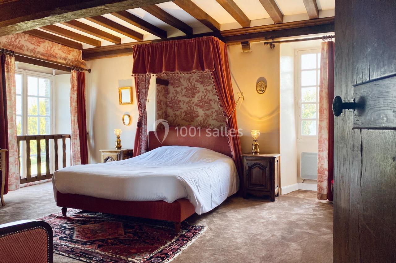 Chambre lumineuse avec lit à baldaquin, tapis rouge, poutres apparentes et fenêtres donnant sur un extérieur verdoyant.