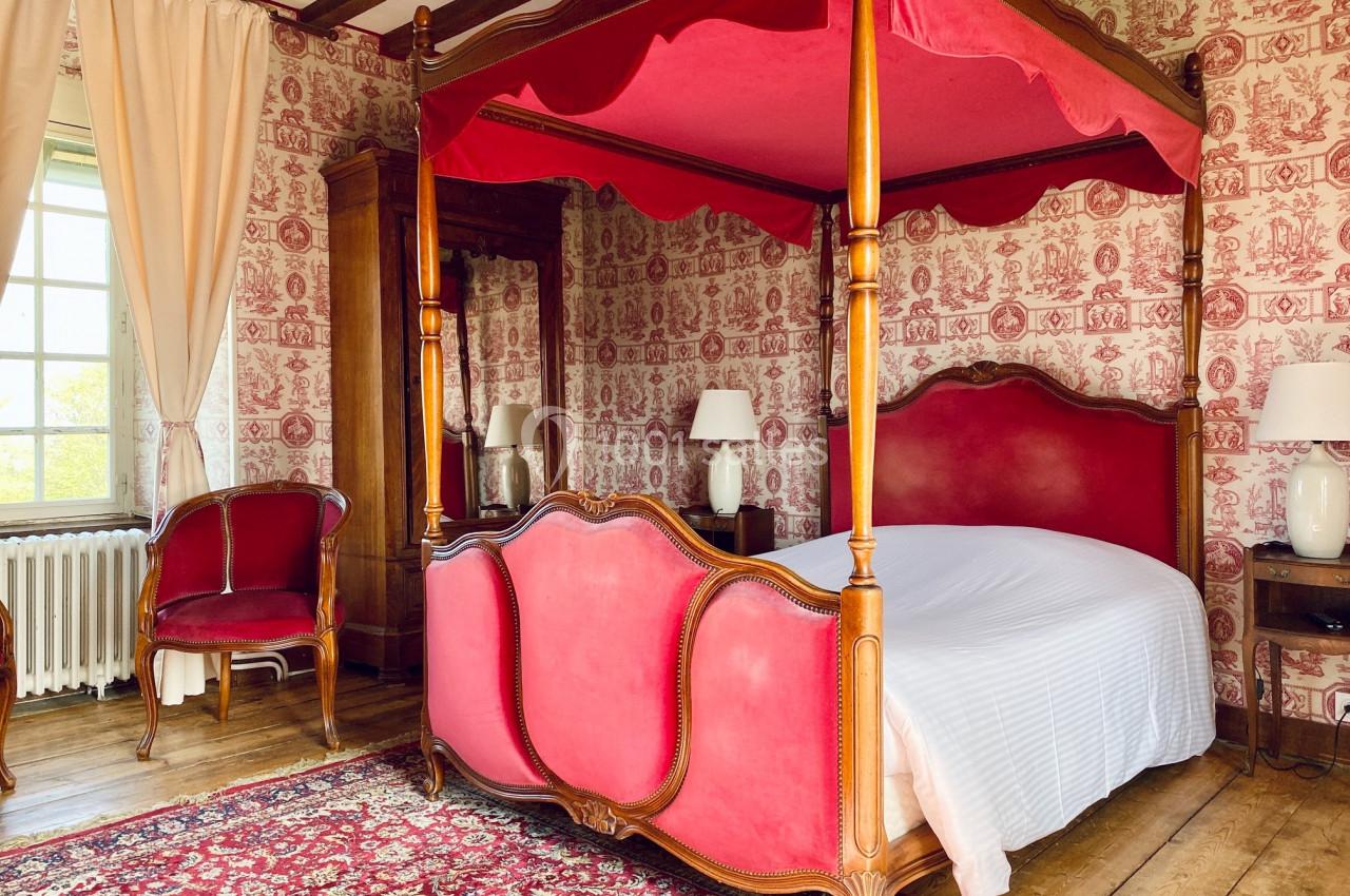 Lit à baldaquin rouge dans une chambre au style ancien avec papier peint à motifs, tapis et mobilier en bois.