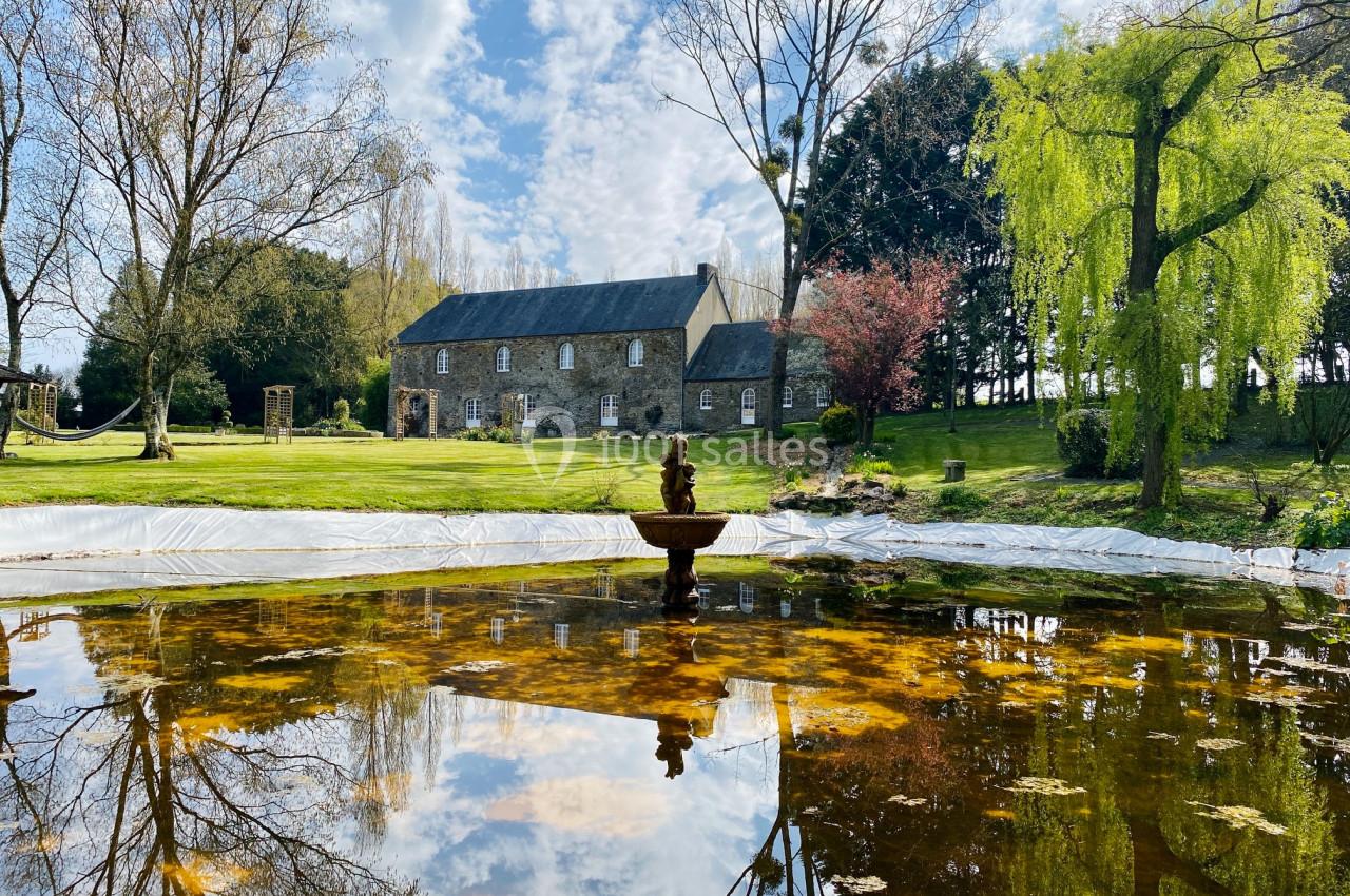 Vue d'une maison en pierre entourée d'arbres, avec un bassin réfléchissant le ciel et un jardin verdoyant.