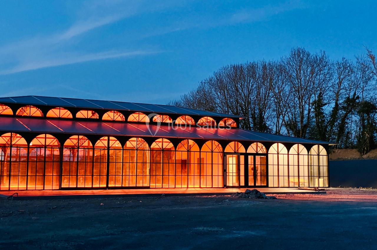 Bâtiment vitré illuminé en orange au crépuscule, entouré d'arbres et d'un sol dégagé.