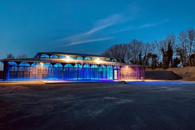 Structure en verre illuminée par des lumières violettes au crépuscule, avec un ciel dégagé en arrière-plan.