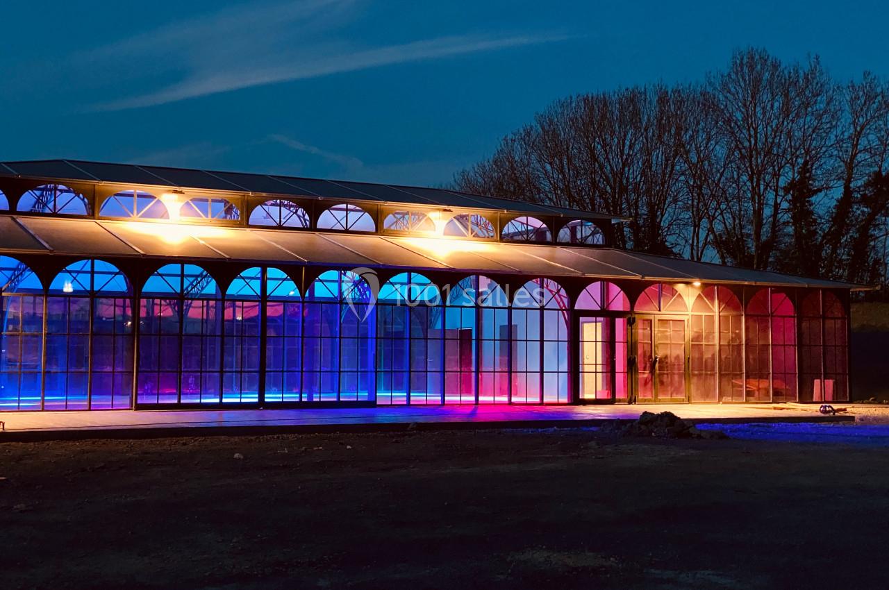 Bâtiment en verre éclairé par des lumières colorées, avec un ciel nocturne et des arbres en arrière-plan.