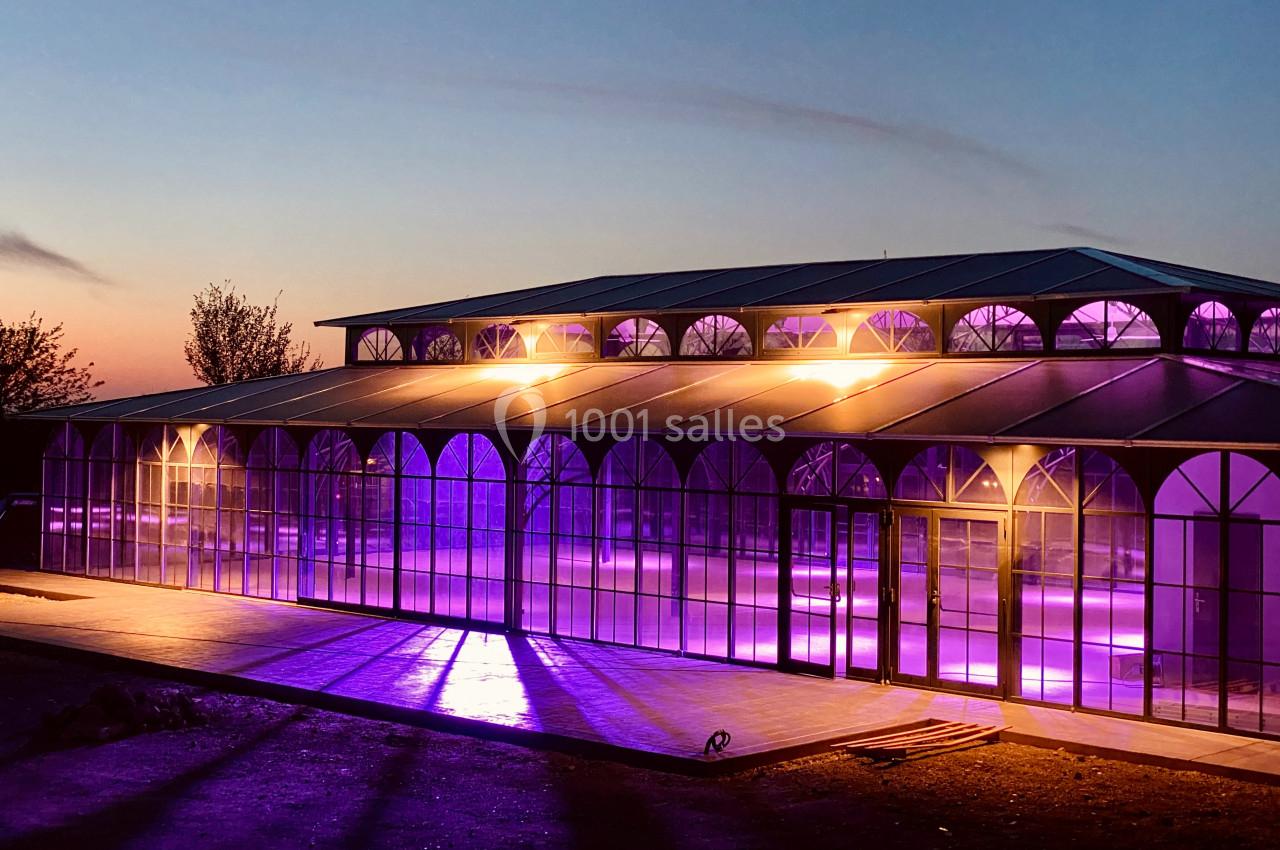Structure en verre illuminée par des lumières violettes au crépuscule, avec un ciel dégagé en arrière-plan.