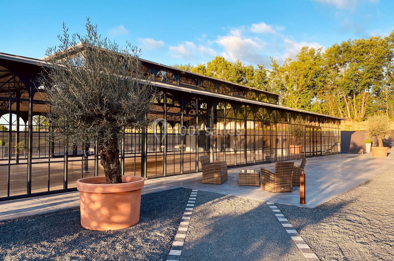 Serre moderne en verre avec mobilier d'extérieur, entourée de végétation et éclairée par une lumière naturelle.