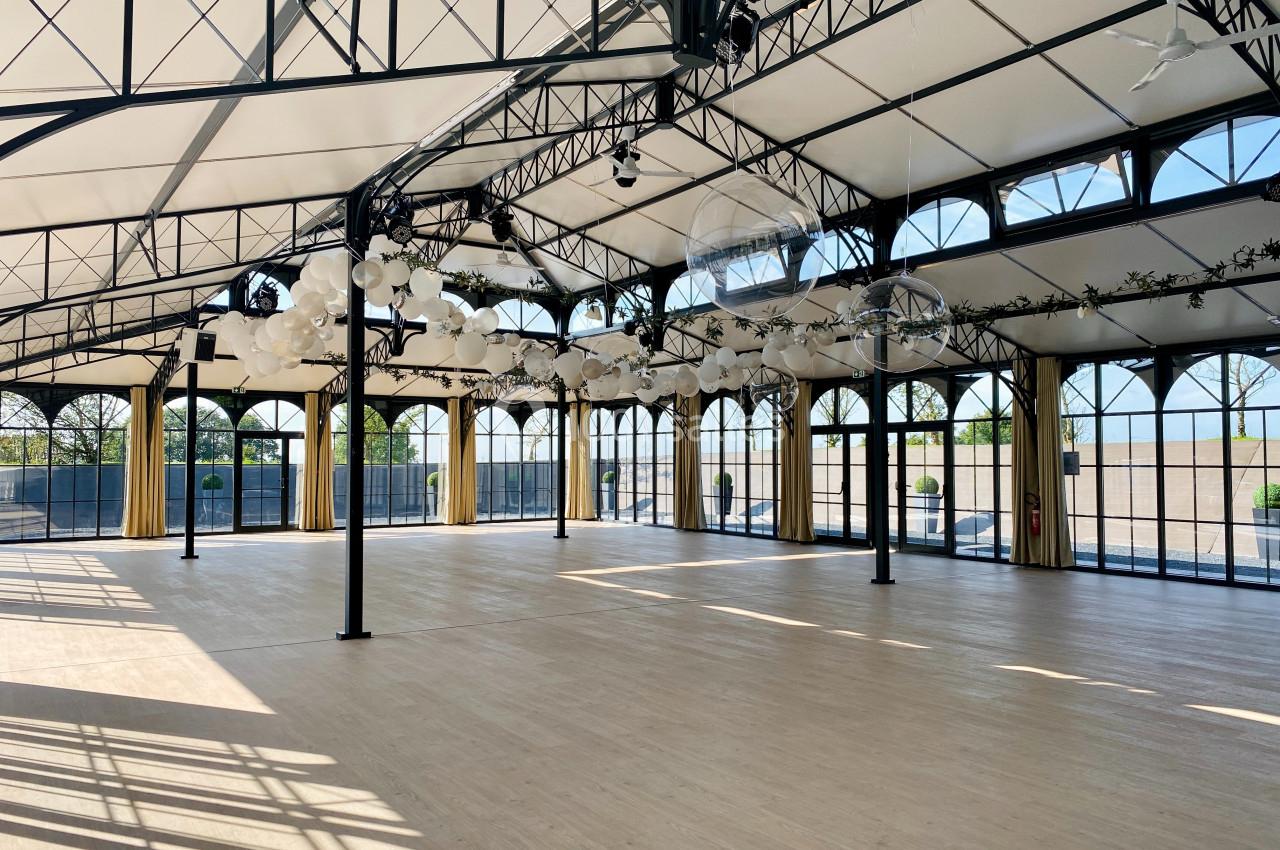 Salle spacieuse avec toit en verrière, structures métalliques noires et décorations suspendues, baignée de lumière naturelle.