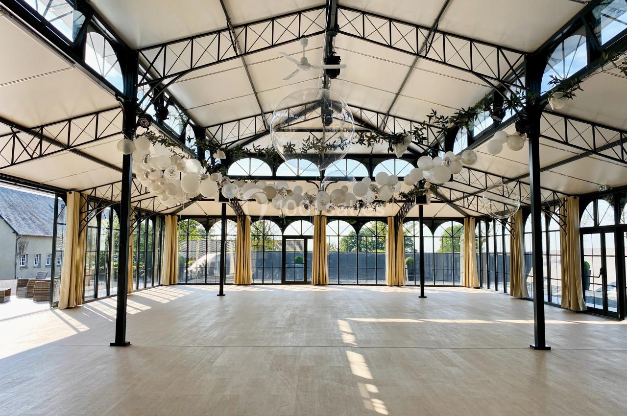 Grande salle lumineuse avec structure métallique, toit vitré, rideaux beiges et guirlandes de lampions suspendus.