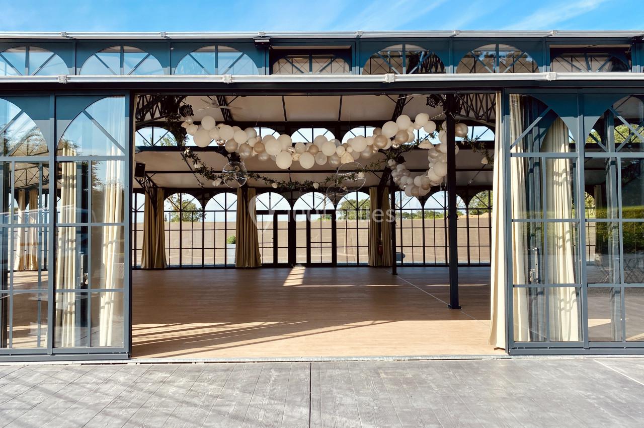 Salle vitrée avec structure métallique, décorée de suspensions lumineuses, donnant sur un espace extérieur dégagé.