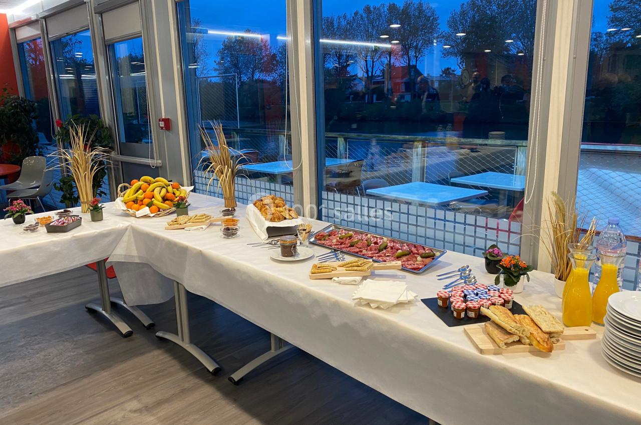 Buffet dressé avec fruits, viennoiseries, jus et mets variés, dans une salle lumineuse avec vue sur l'extérieur.