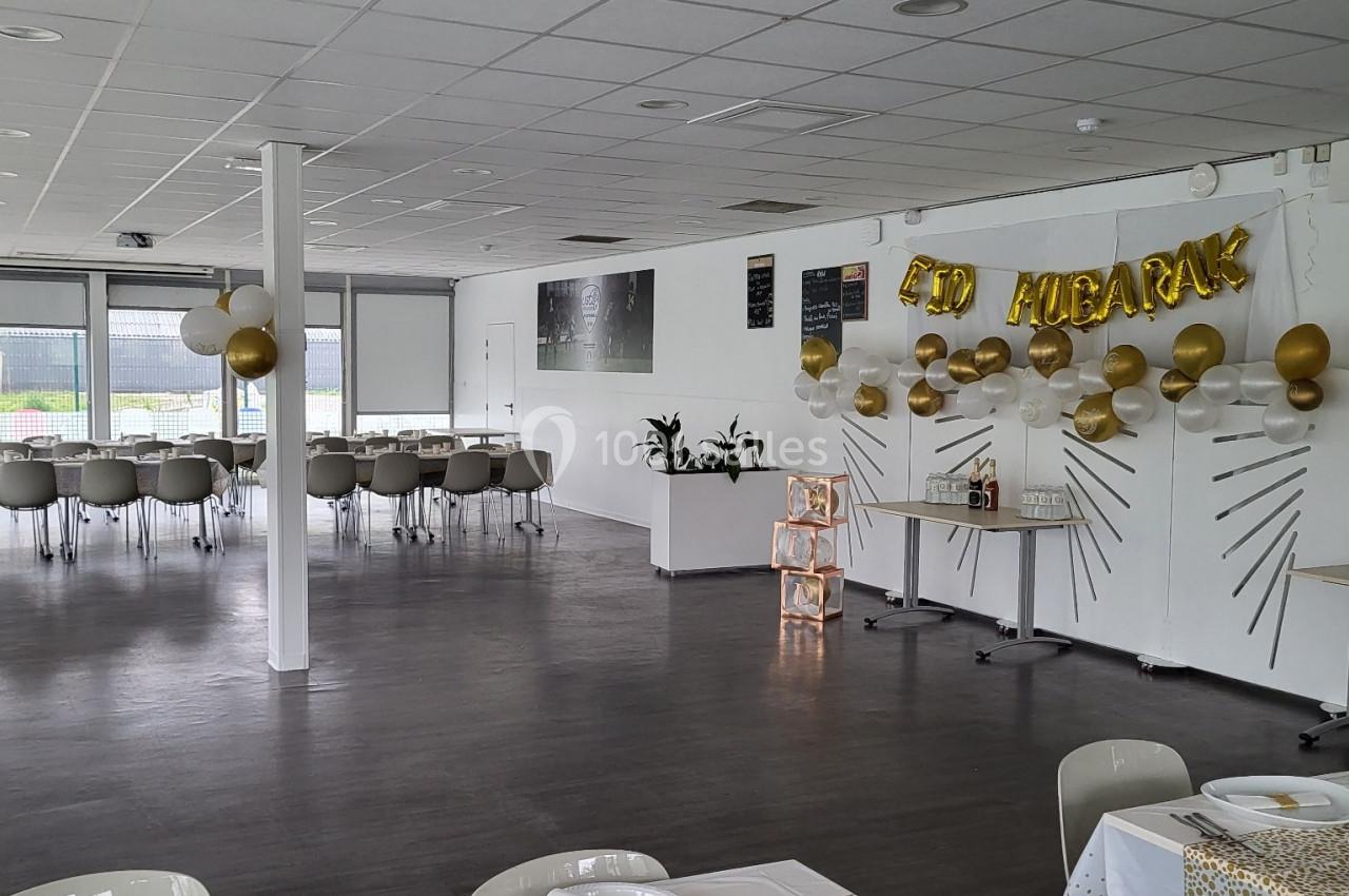 Salle décorée pour une fête avec des tables dressées, des ballons dorés et une bannière ’Eid Mubarak’.