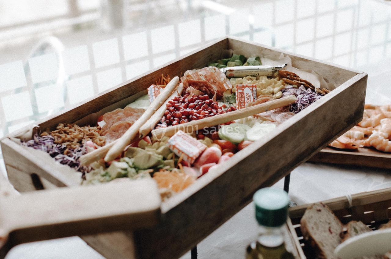 Plateau en bois rempli de légumes frais, fruits, charcuterie et grissini, présenté sur une table.