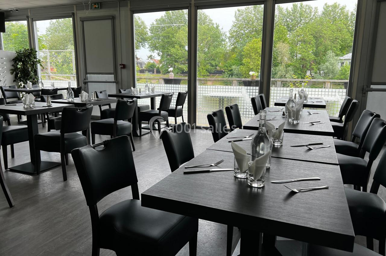 Salle de restaurant lumineuse avec tables dressées, chaises noires et vue sur un espace extérieur verdoyant.