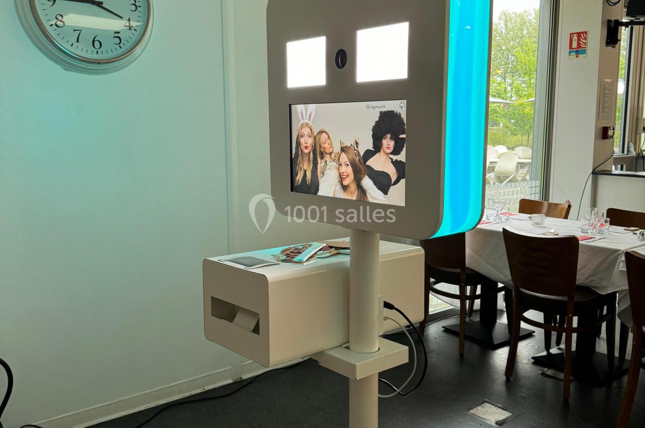 Borne photo interactive avec écran lumineux affichant des photos, installée dans une salle avec une horloge murale.