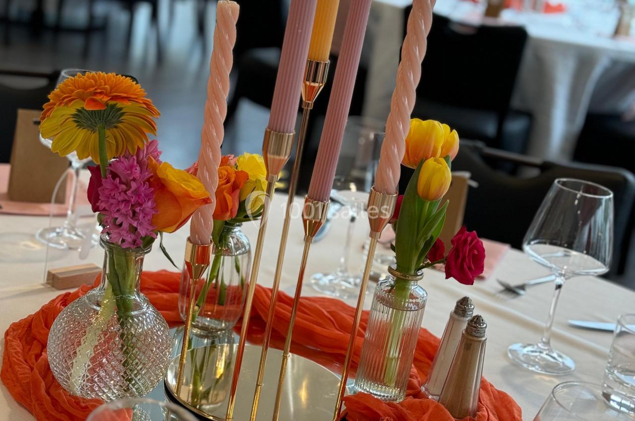 Centre de table avec bougies torsadées, fleurs colorées et nappe orange, entouré de verres et couverts.
