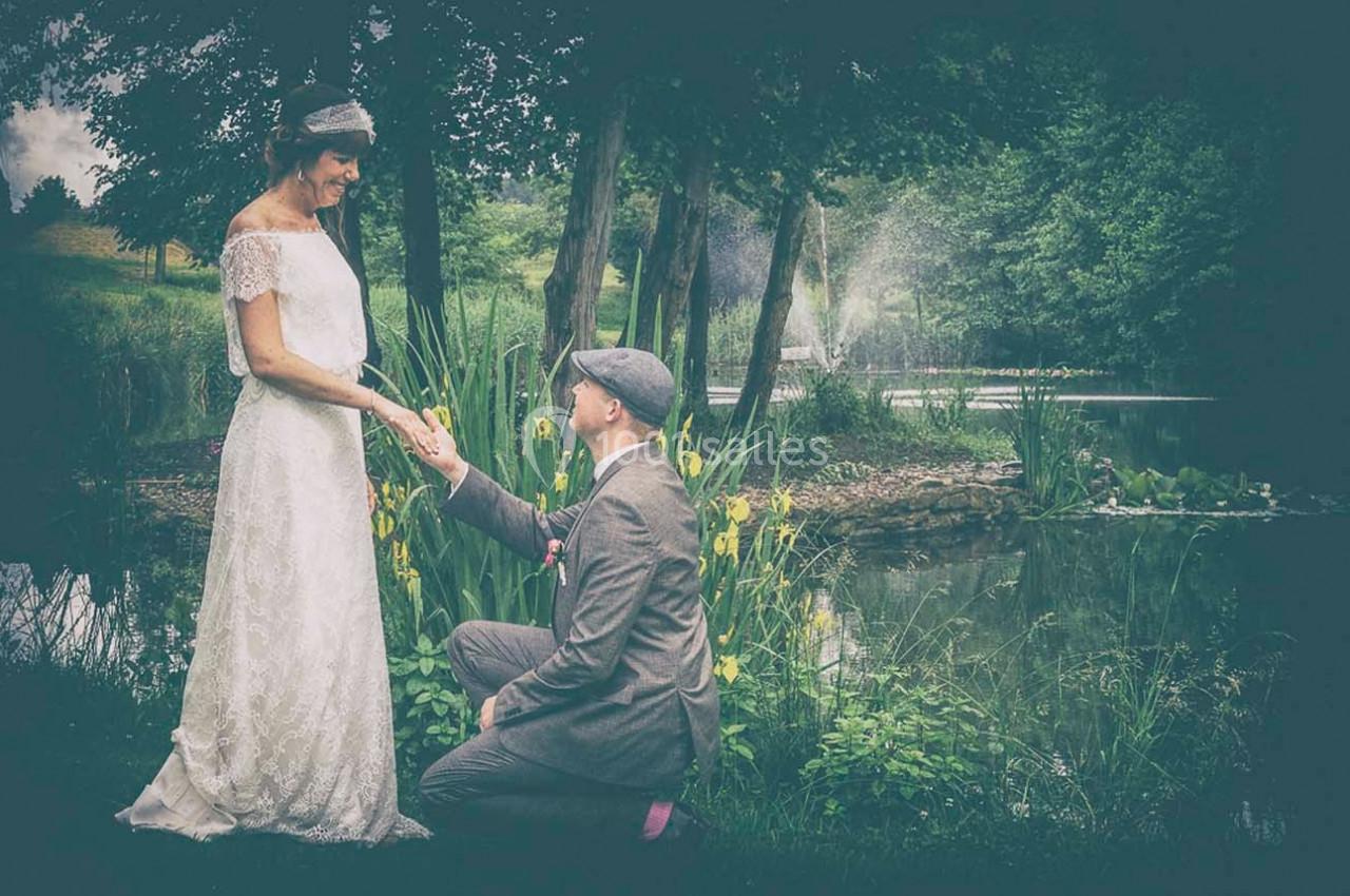 Un homme à genoux propose à une femme en robe blanche près d'un étang entouré de végétation.