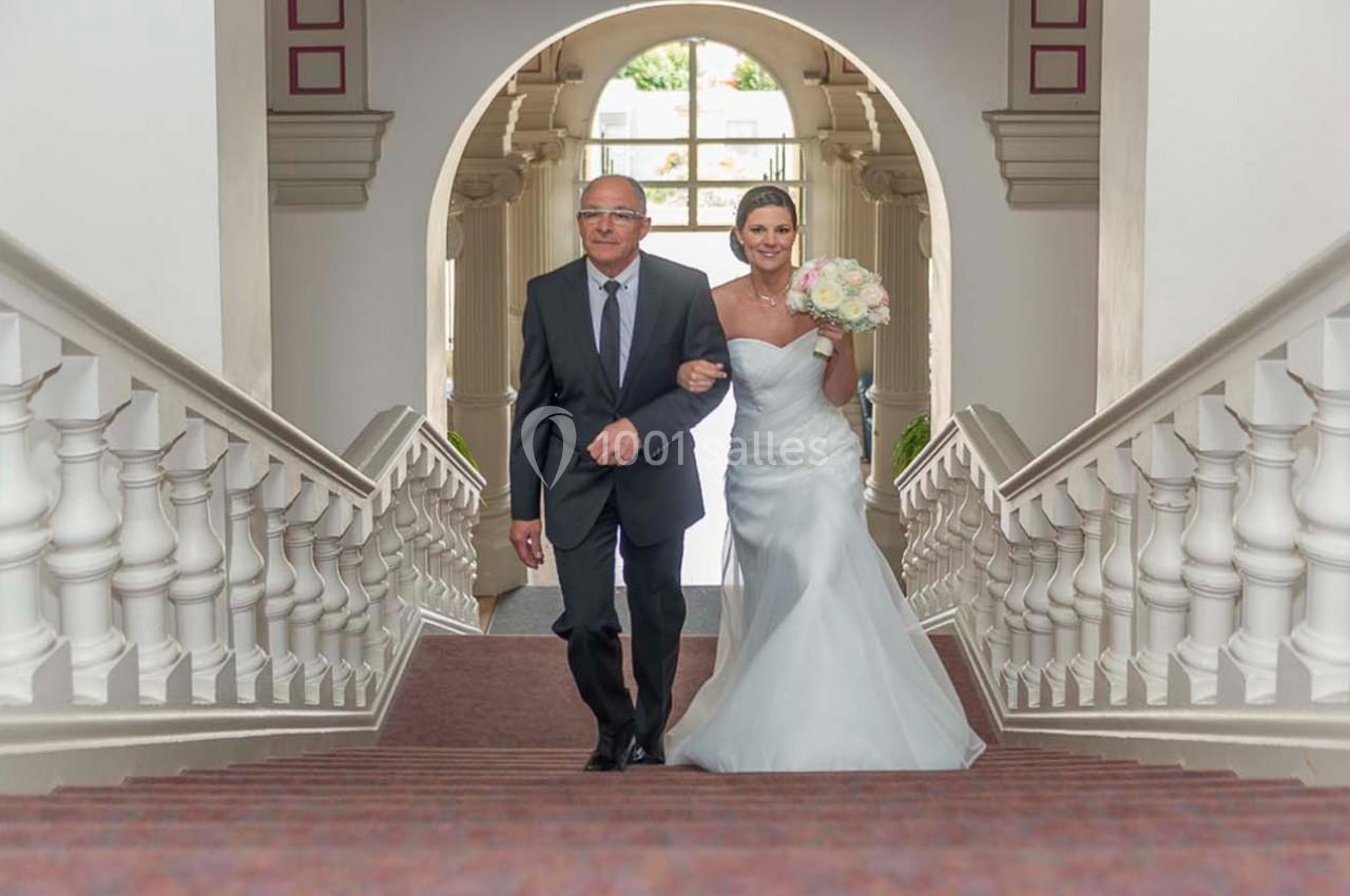 Un homme en costume accompagne une mariée en robe blanche montant un escalier dans un bâtiment lumineux.