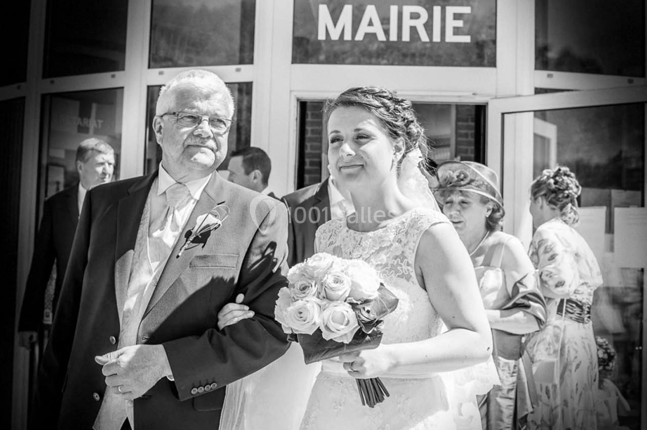Une mariée souriante tenant un bouquet de roses blanches sort de la mairie au bras d'un homme en costume.