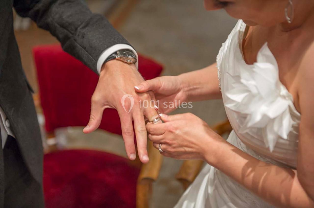 Une mariée passe une alliance au doigt de son partenaire, assis sur une chaise rouge.