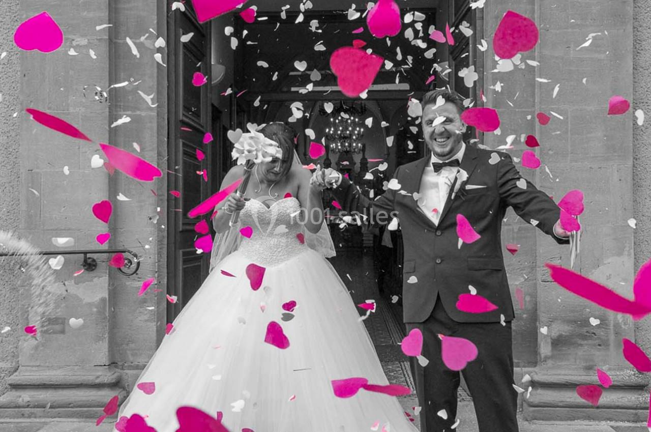 Un couple souriant en tenue de mariage sort d'un bâtiment sous une pluie de confettis roses et blancs.