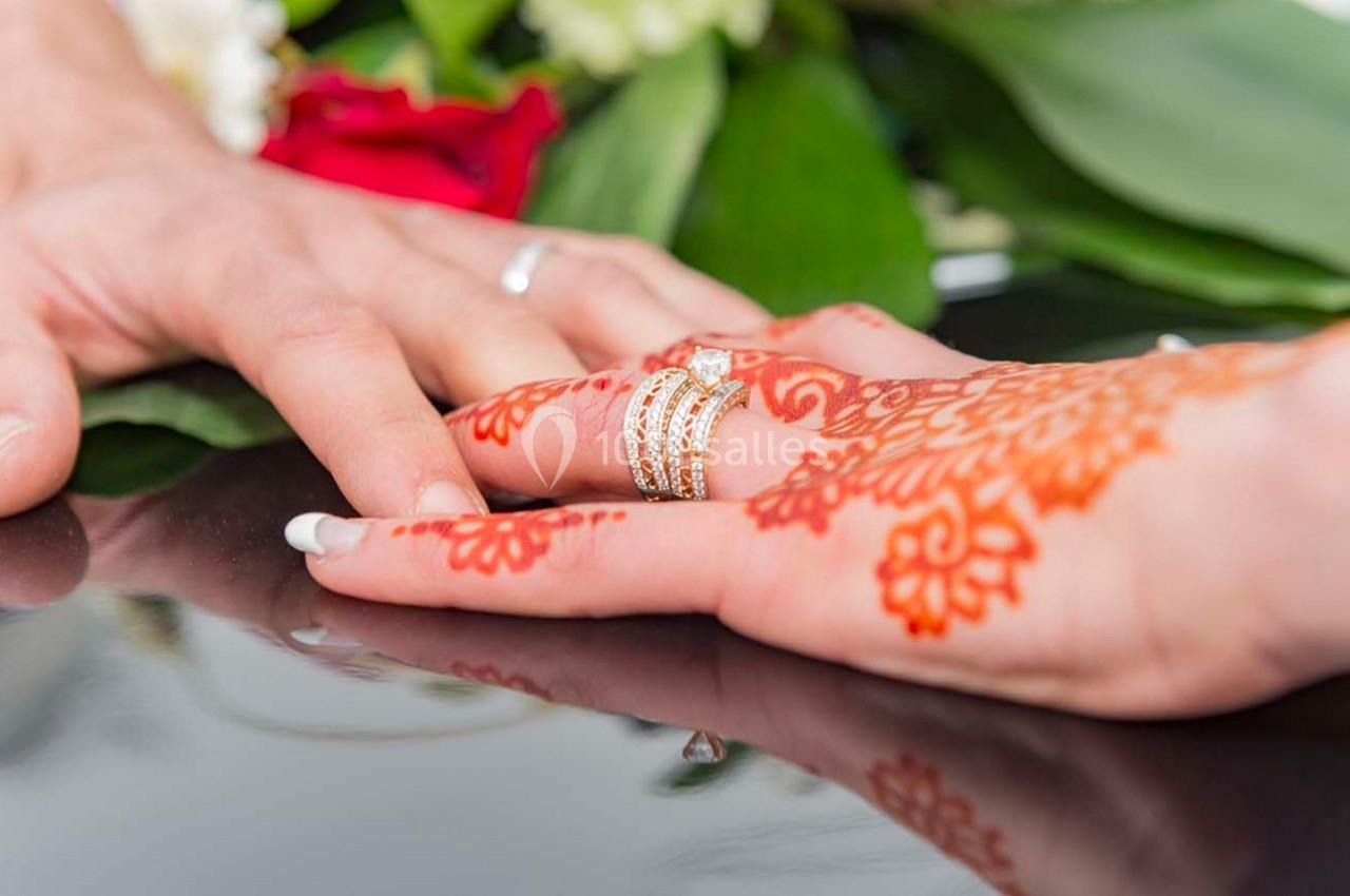 Main ornée de henné et bagues touchant une autre main sur une surface réfléchissante, avec des fleurs en arrière-plan.