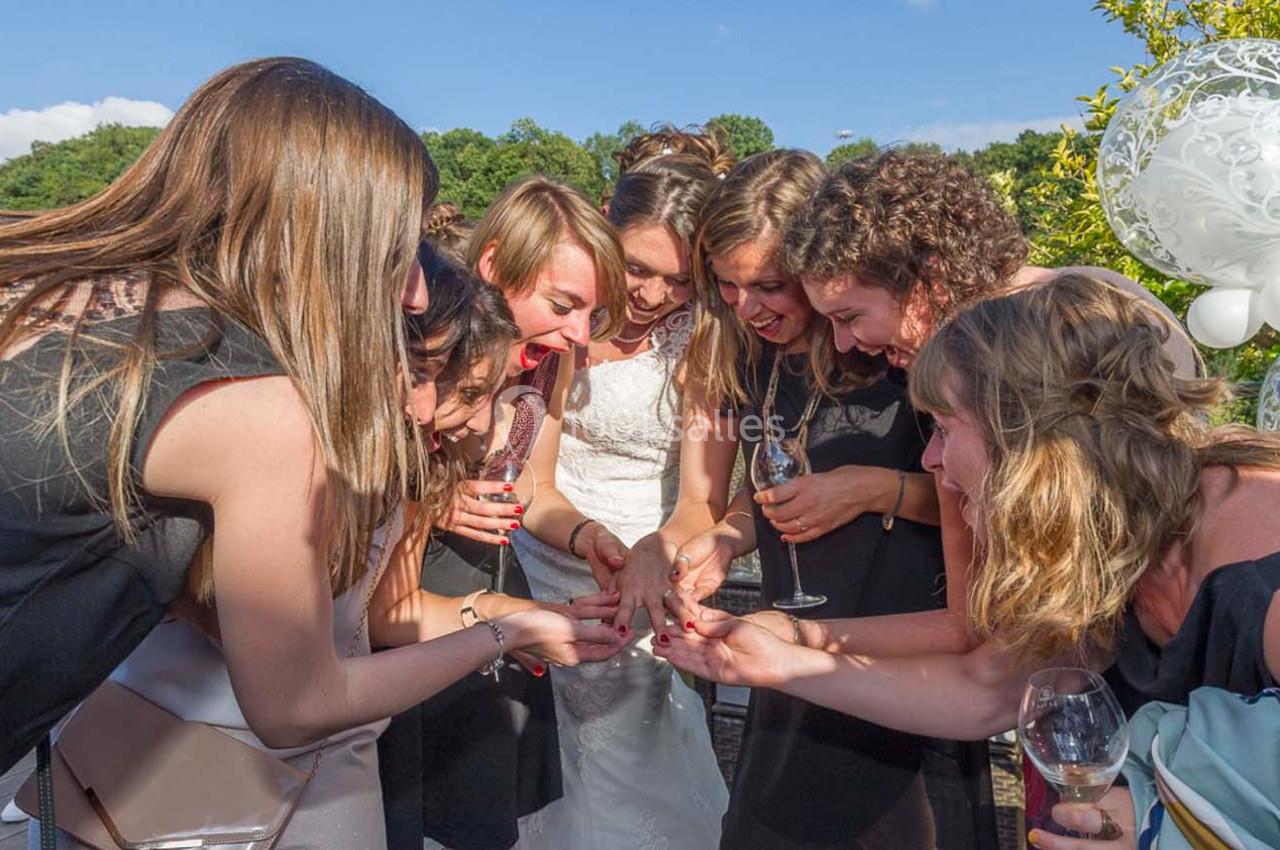Un groupe de femmes souriantes entoure une mariée en regardant sa main avec enthousiasme, en extérieur.