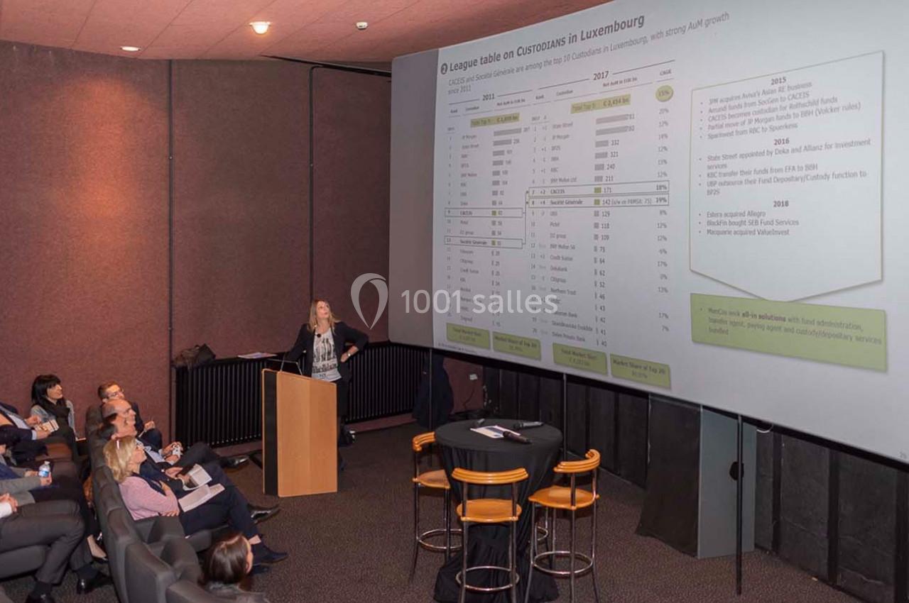 Une conférencière présente un tableau de données projeté sur un écran à un public assis dans une salle sombre.