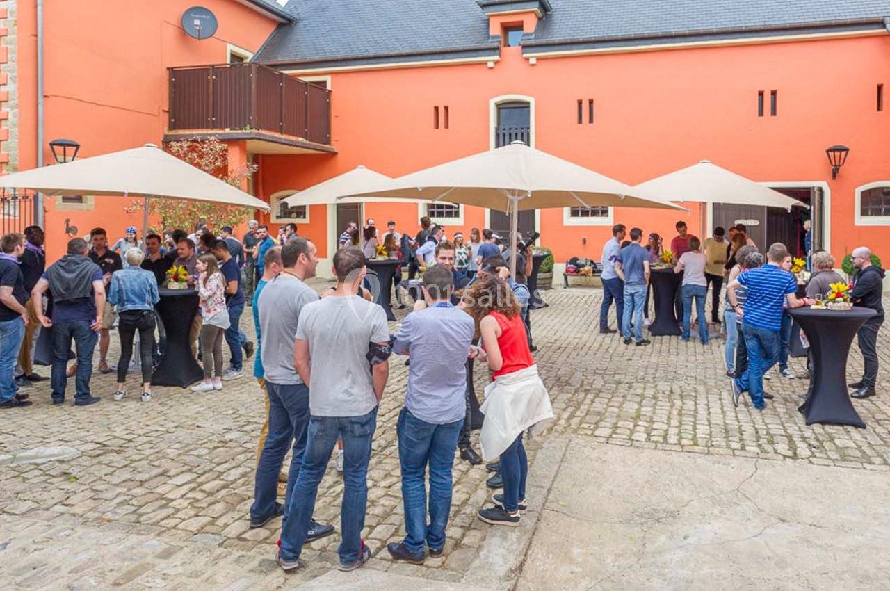 Groupe de personnes rassemblées dans une cour pavée devant un bâtiment orange, discutant sous des parasols.