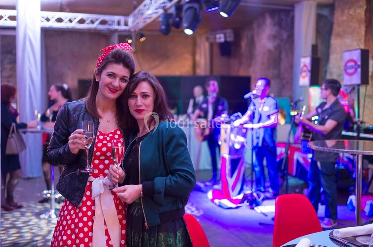 Deux femmes souriantes tenant des verres de champagne dans une salle animée avec un groupe jouant en arrière-plan.
