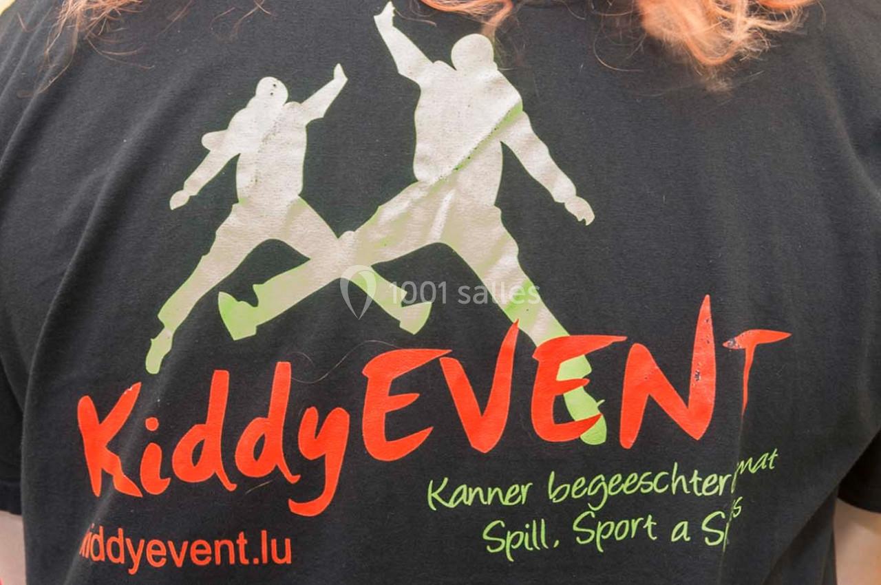 Dos d’un t-shirt noir avec un logo représentant deux silhouettes sautant et des inscriptions colorées en luxembourgeois.