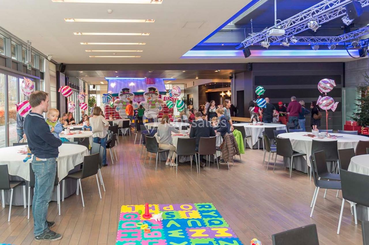 Salle décorée avec des ballons colorés, des tables rondes et des familles participant à un événement festif.