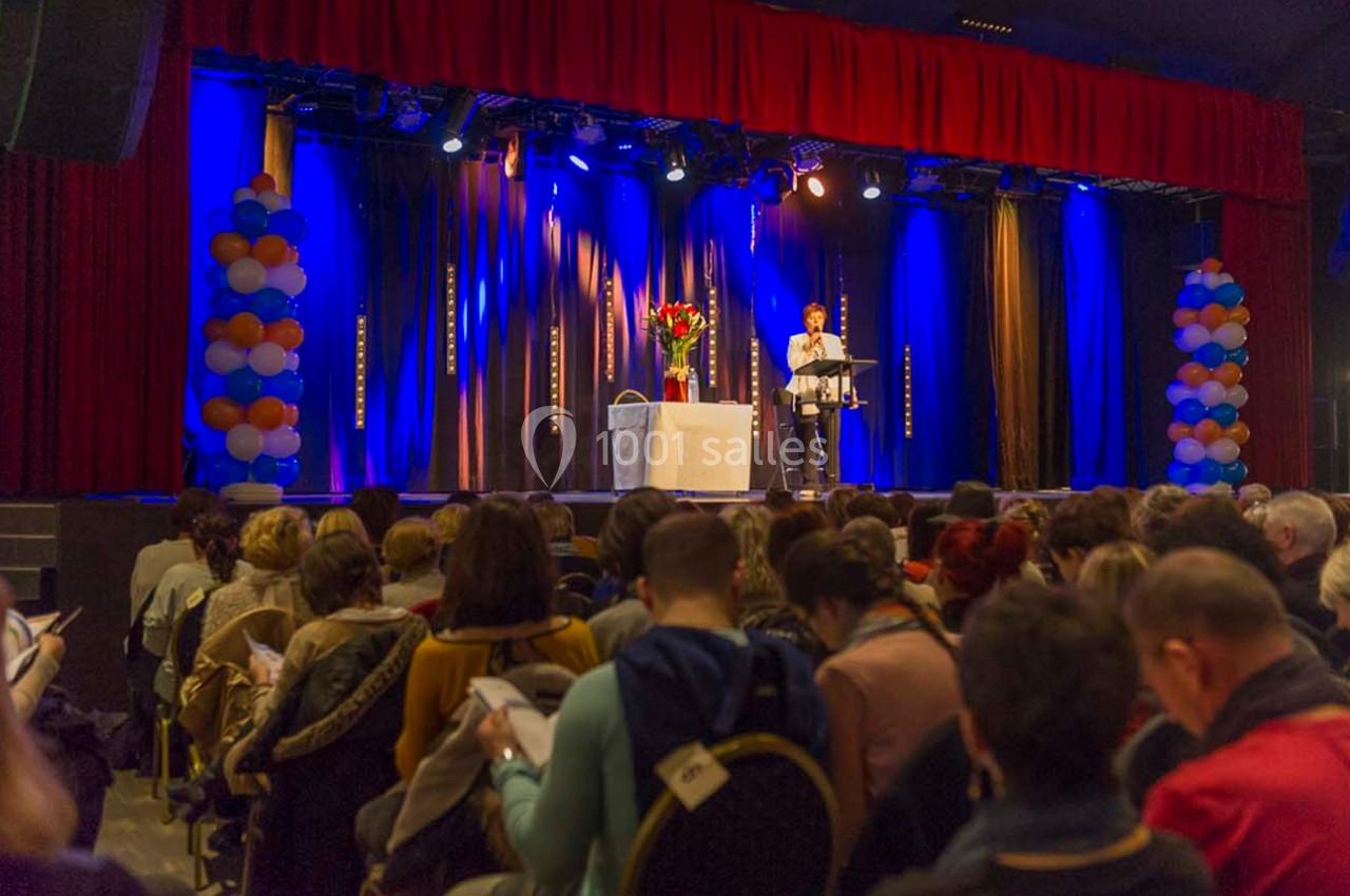 Une personne parle sur scène devant un public assis, avec des rideaux bleus et rouges et des ballons en arrière-plan.