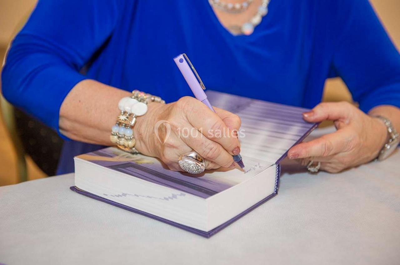 Une personne signe un livre avec un stylo violet, portant des bijoux et une tenue bleue.