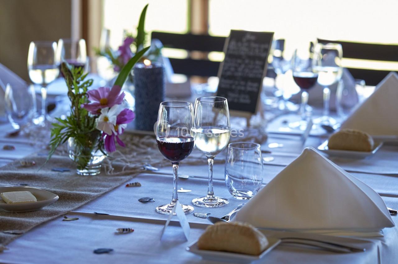 Table dressée avec nappes blanches, verres de vin, fleurs colorées et menu sur ardoise en arrière-plan.