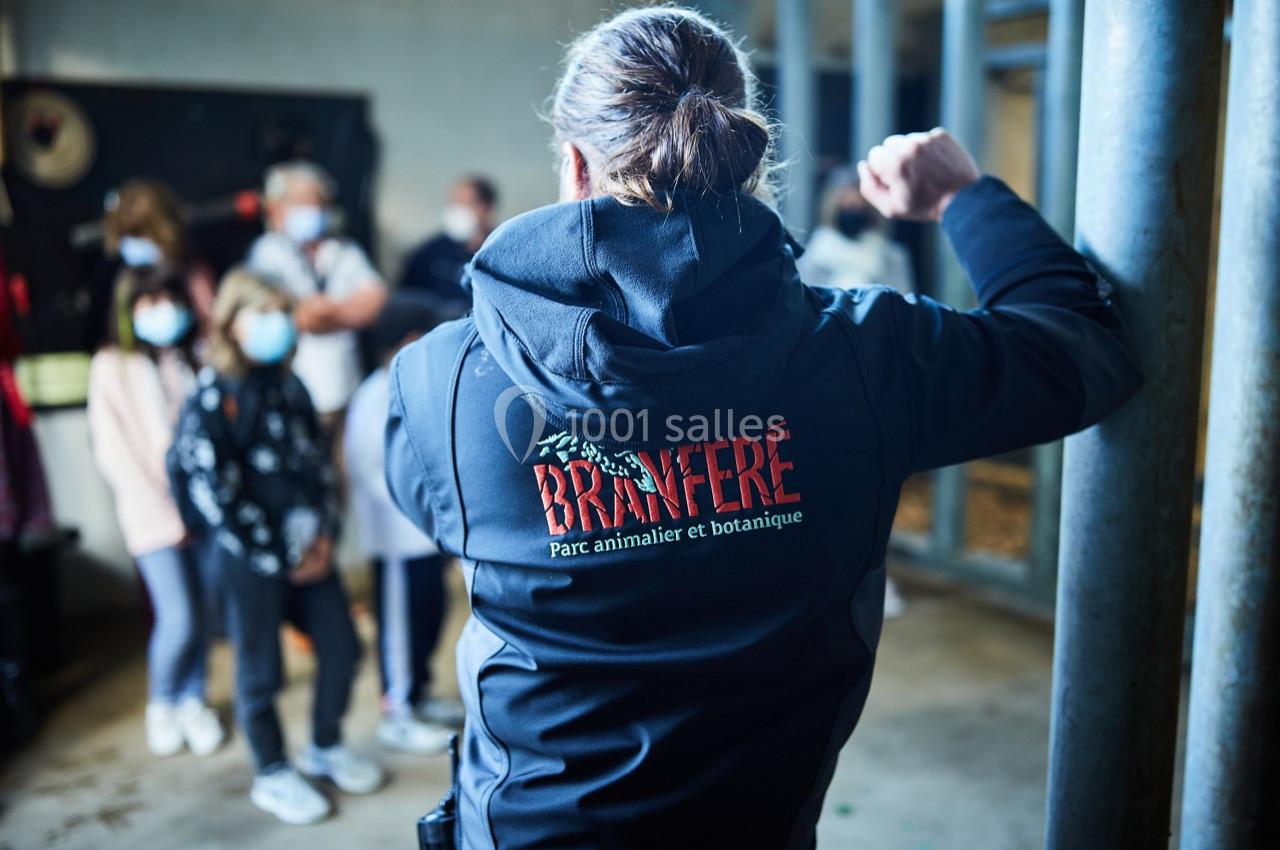 Une personne de dos portant une veste ’Branféré’ devant un groupe de visiteurs dans un espace intérieur.